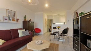 Ein modernes Wohnzimmer mit einem roten Sofa, einem Couchtisch und einem Essbereich mit weißem Tisch und Stühlen. Es gibt eine Küchenzeile mit Schränken und einem Kühlschrank.
