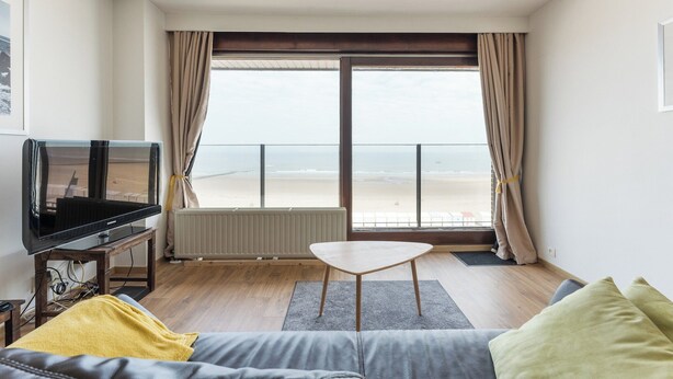 A living room with a TV, a sofa, a coffee table, and a view of the beach through large windows.