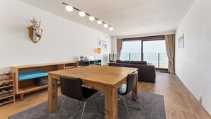 A modern living room with a wooden dining table, black chairs, a sofa, a mounted deer head, and a large window with a view.