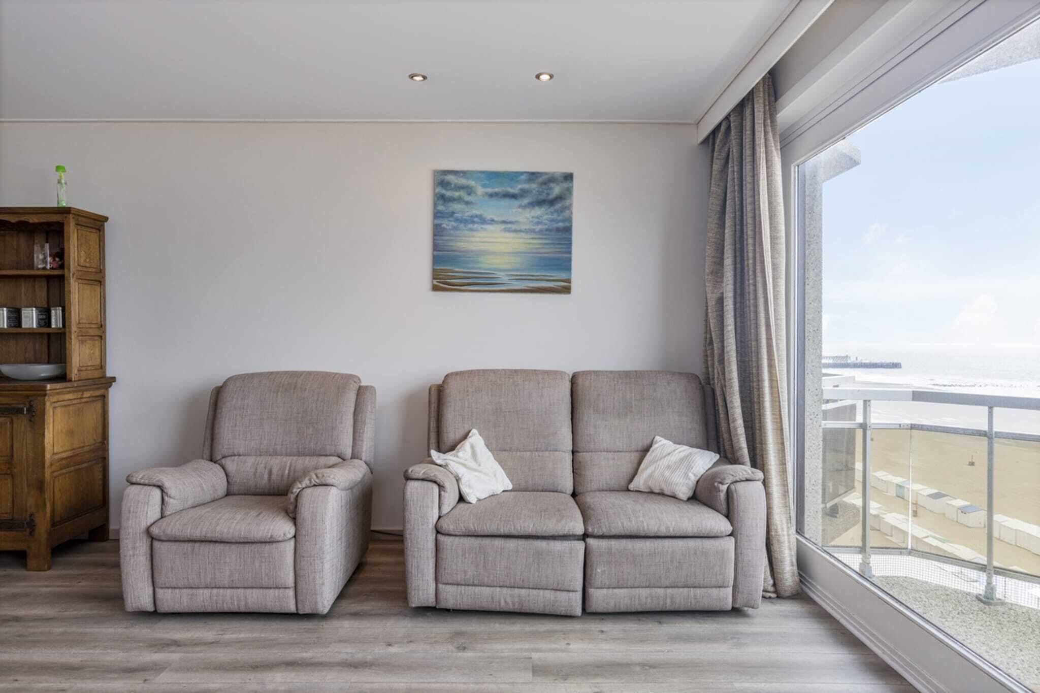 A room with two grey armchairs, a wooden cabinet, a painting of a sunset, and a large window with a view of the beach.
