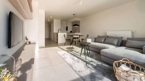 A modern living room with a grey sofa, a dining table with chairs, and a TV mounted on the wall.