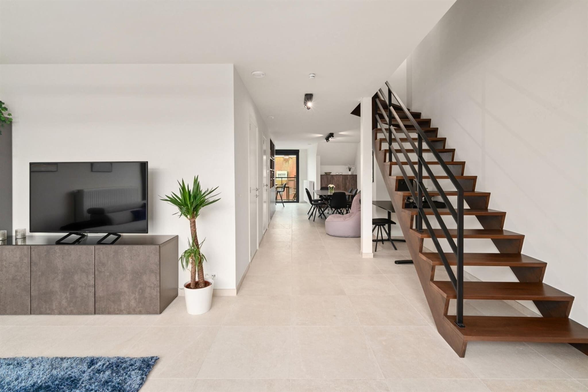 A modern living room with a flat-screen TV, a grey cabinet, a potted plant, and a staircase with wooden steps and black railings.