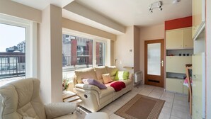 A living room with a sofa, a chair, a radiator, a window, and a kitchen area with cabinets. - Flat in Bruges with Panoramic City Views (BLANKENBERGE)