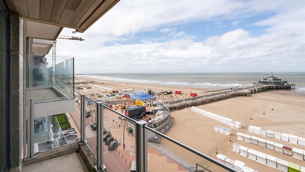 Blick auf einen Strand vom Balkon mit Glasgeländer, einen Steg und Strandstühle.