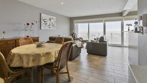 A spacious living area with a dining table, chairs, and a sofa. There is a large window providing a view of the outdoors. - Apartment in Blankenberge by the Marina (BLANKENBERGE)
