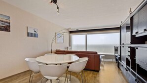 A modern living room with a round dining table, white chairs, a brown sofa, and a large window offering a view of the sea.