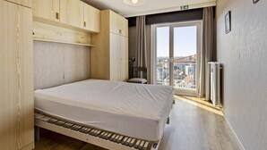 A compact bedroom with a bed, wooden wardrobe, and a view of a cityscape through a large window.