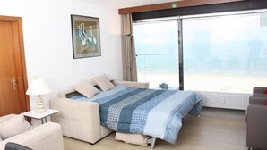 Une chambre d’hôtel avec un lit, une chaise, une lampe, un tableau au mur et une grande fenêtre donnant sur la plage.
