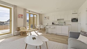 A modern living room with a white sofa, a dining table, and a kitchen area with white cabinets and a built-in oven.