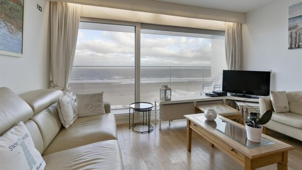 Una sala de estar moderna con un amplio ventanal con vistas a la playa, un televisor de pantalla plana y un rincón de estar confortable.