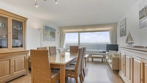 Amplia sala de estar con un gran ventanal que ofrece vista al mar, una mesa de comedor con sillas de mimbre, un televisor y un mueble con puertas de cristal.
