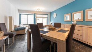 A modern living room with a dining area, featuring a large window, a sofa, a coffee table, and a wooden dining table with chairs.