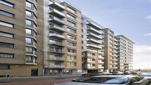 A multi-story residential building with balconies, a parking area, and a clear sky.