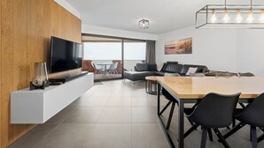 A modern living room with a flat-screen TV, wooden panel wall, and a dining area with a wooden table and black chairs. - Apartment in Blankenberge with Sea Views (BLANKENBERGE)
