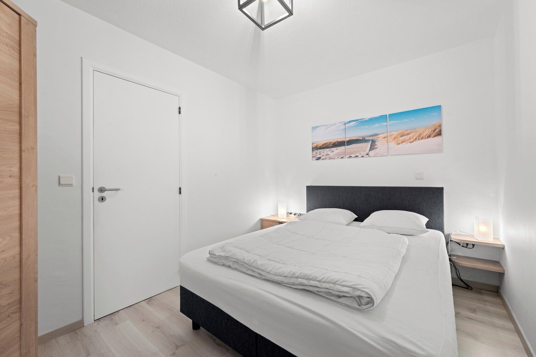 A bedroom with a bed, bedside tables, a nightstand, a door, and a wall-mounted picture.