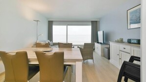Una habitación de hotel moderna con zona de comedor, televisor y un amplio ventanal con cortinas.