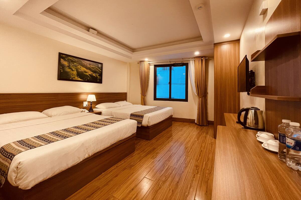 Habitación de hotel con dos camas, piso de madera, un cuadro en la pared, una ventana con cortinas y una cafetera sobre la mesita de noche.