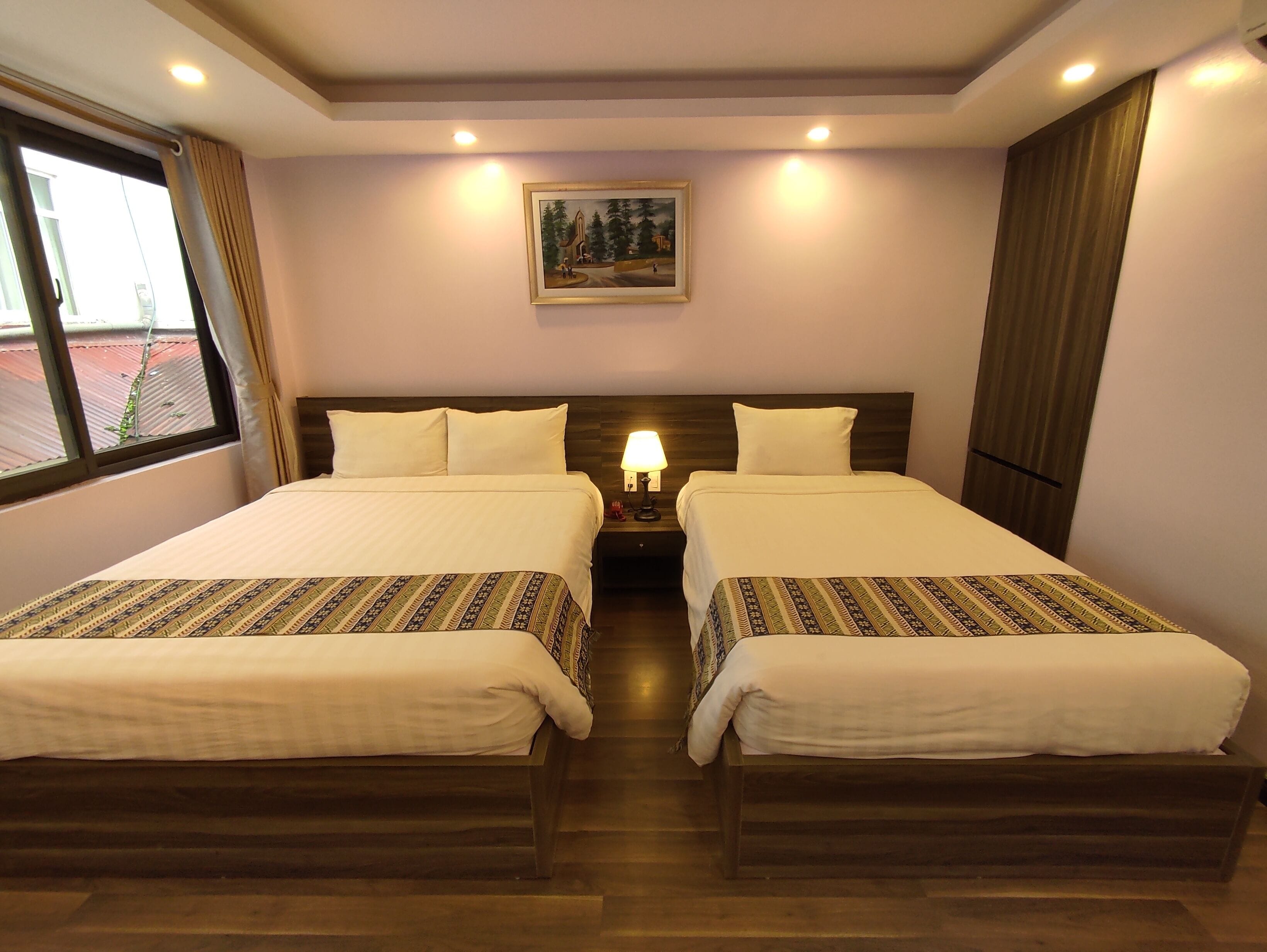A hotel room with two beds, a nightstand with a lamp, a painting on the wall, and a window with curtains.