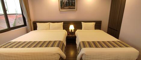 A hotel room with two beds, a nightstand with a lamp, a painting on the wall, and a window with curtains.