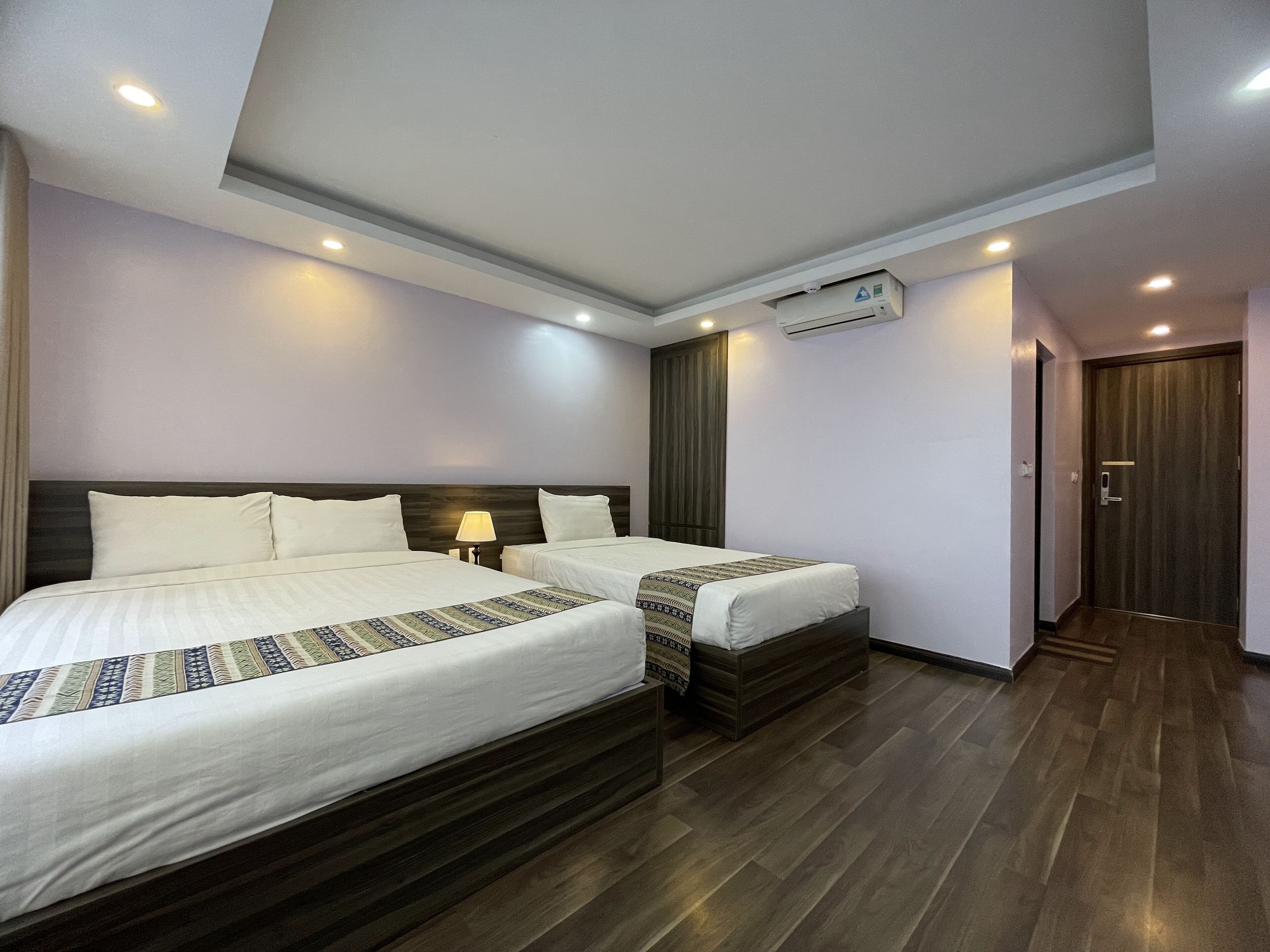 A hotel room with two beds, a wooden headboard, a ceiling light, an air conditioning unit, and a wooden floor.