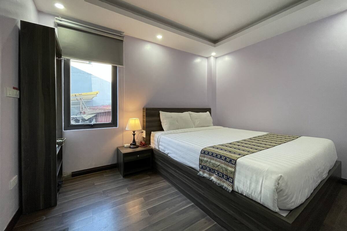 Habitación de hotel moderna con una cama grande, una mesita de noche, una lámpara y una ventana que da a una vista de construcción.