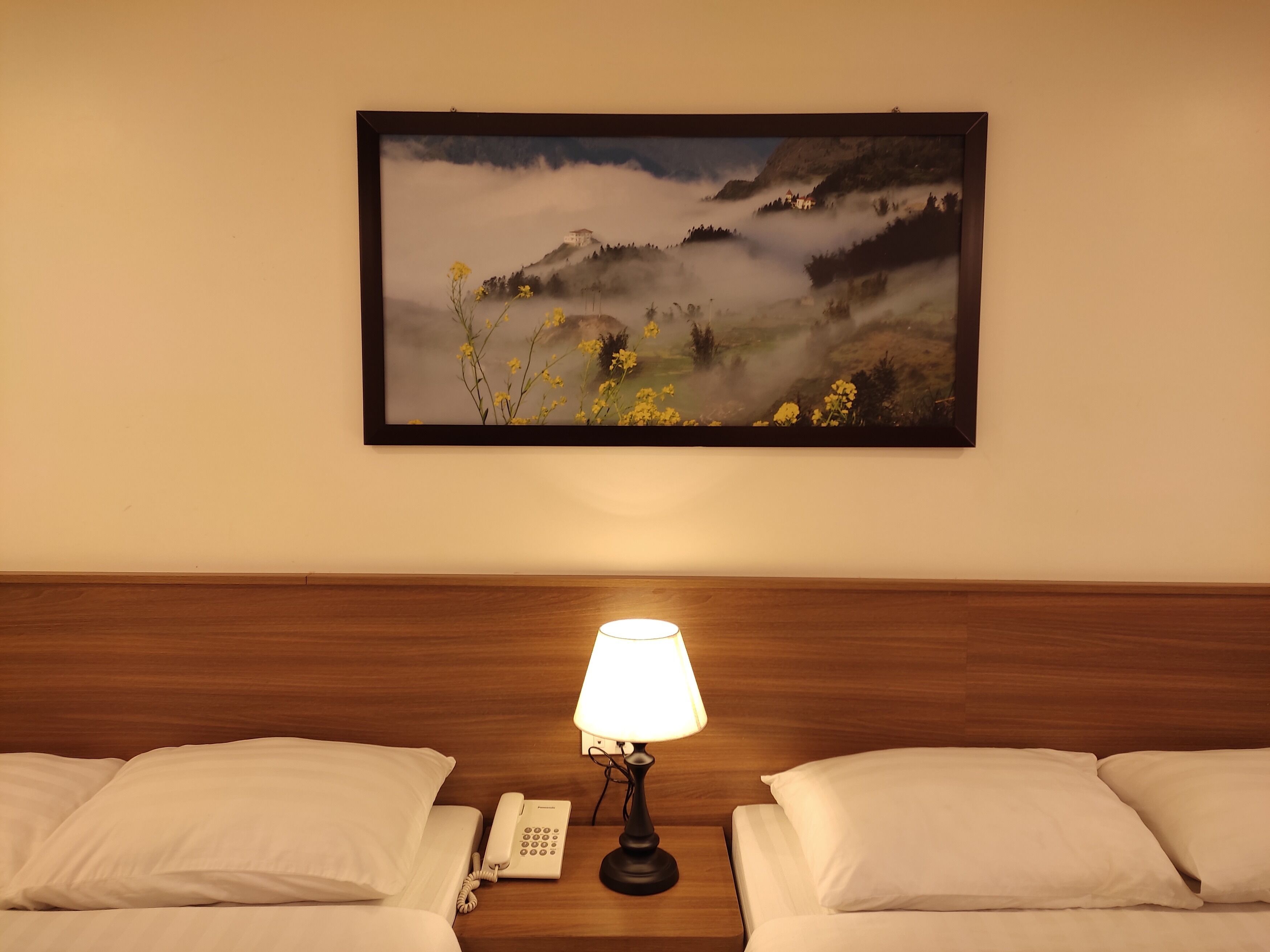 Ein Hotelzimmer mit einem Bett, einer Nachttischlampe, einem Telefon und einem gerahmten Landschaftsbild an der Wand.
