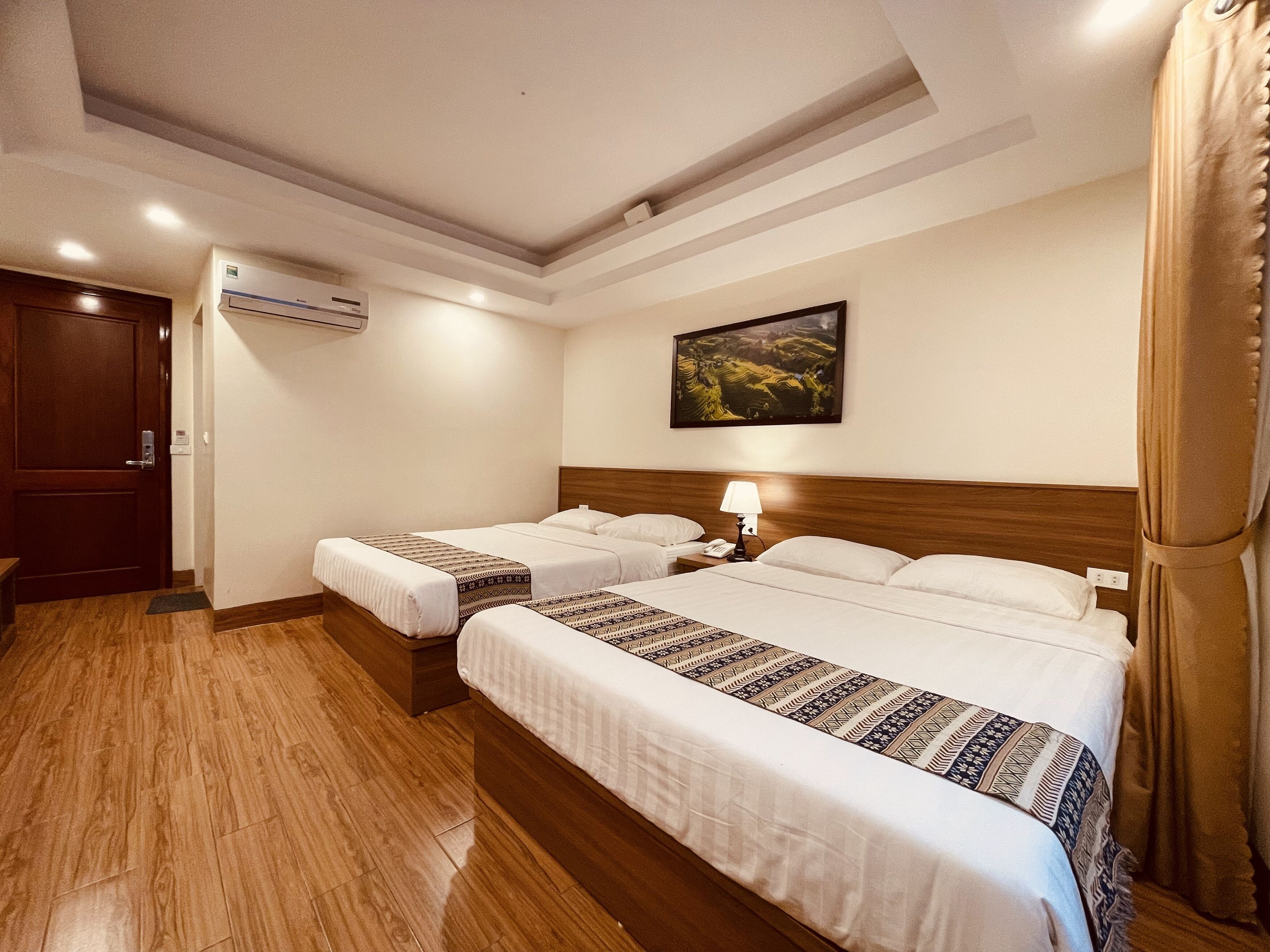 A hotel room with two beds, a wooden floor, an air conditioning unit, a painting on the wall, and a door.