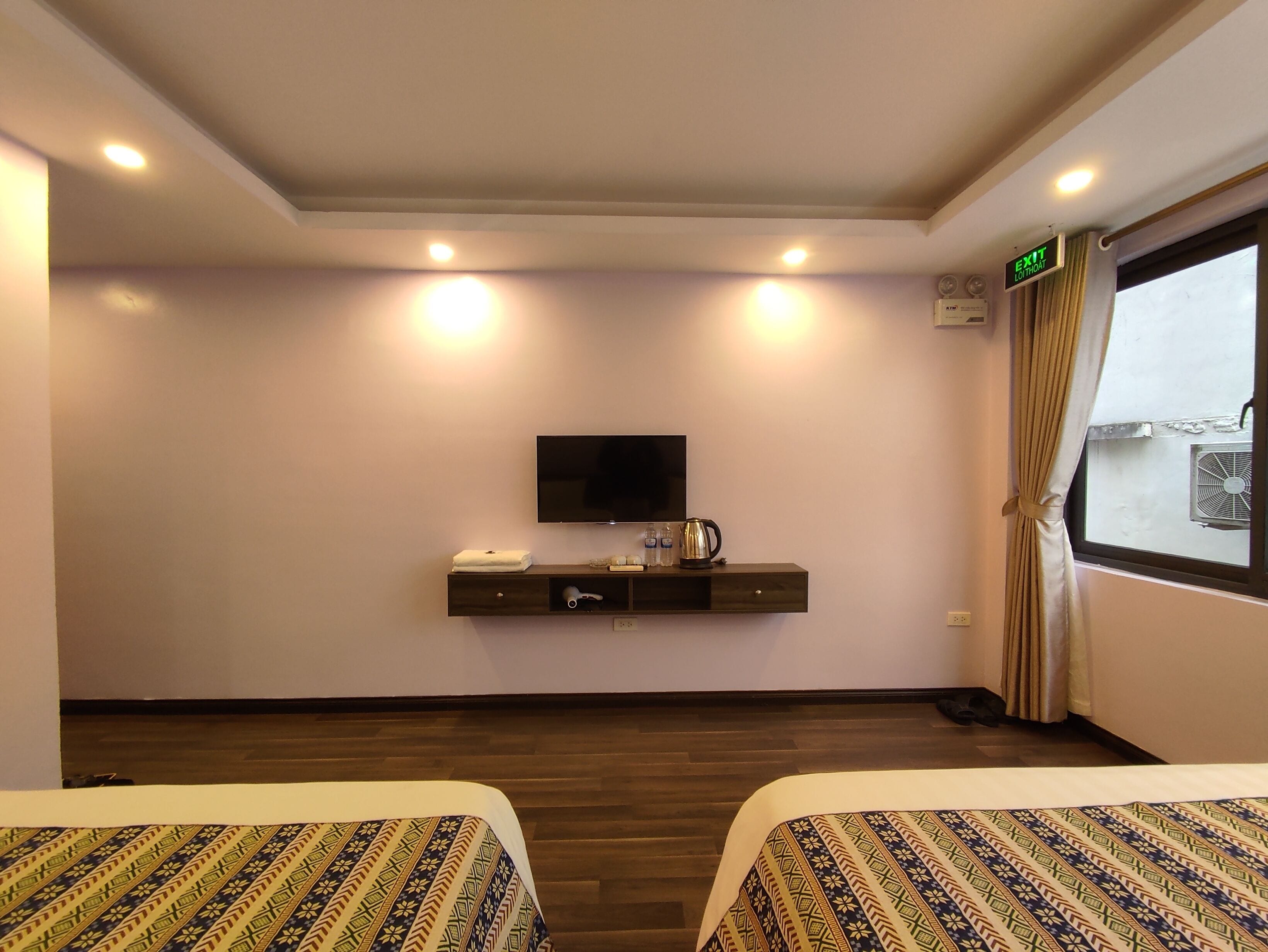 A hotel room with two beds, a flat-screen TV, a small console table with a kettle and glasses, and an exit sign.