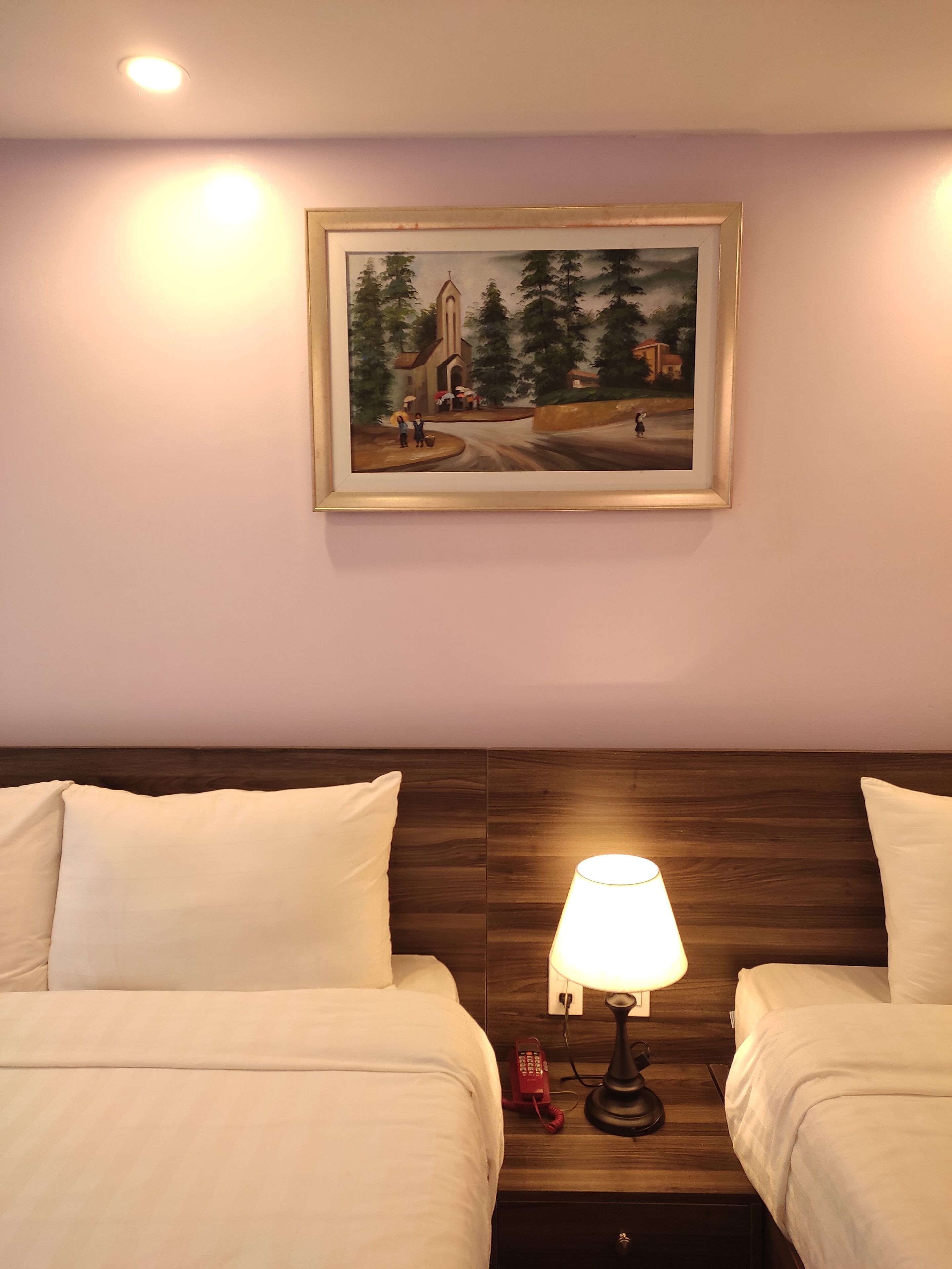 Une chambre d’hôtel avec deux lits, une table de chevet avec une lampe, un tableau représentant un paysage rural et une tête de lit en bois.
