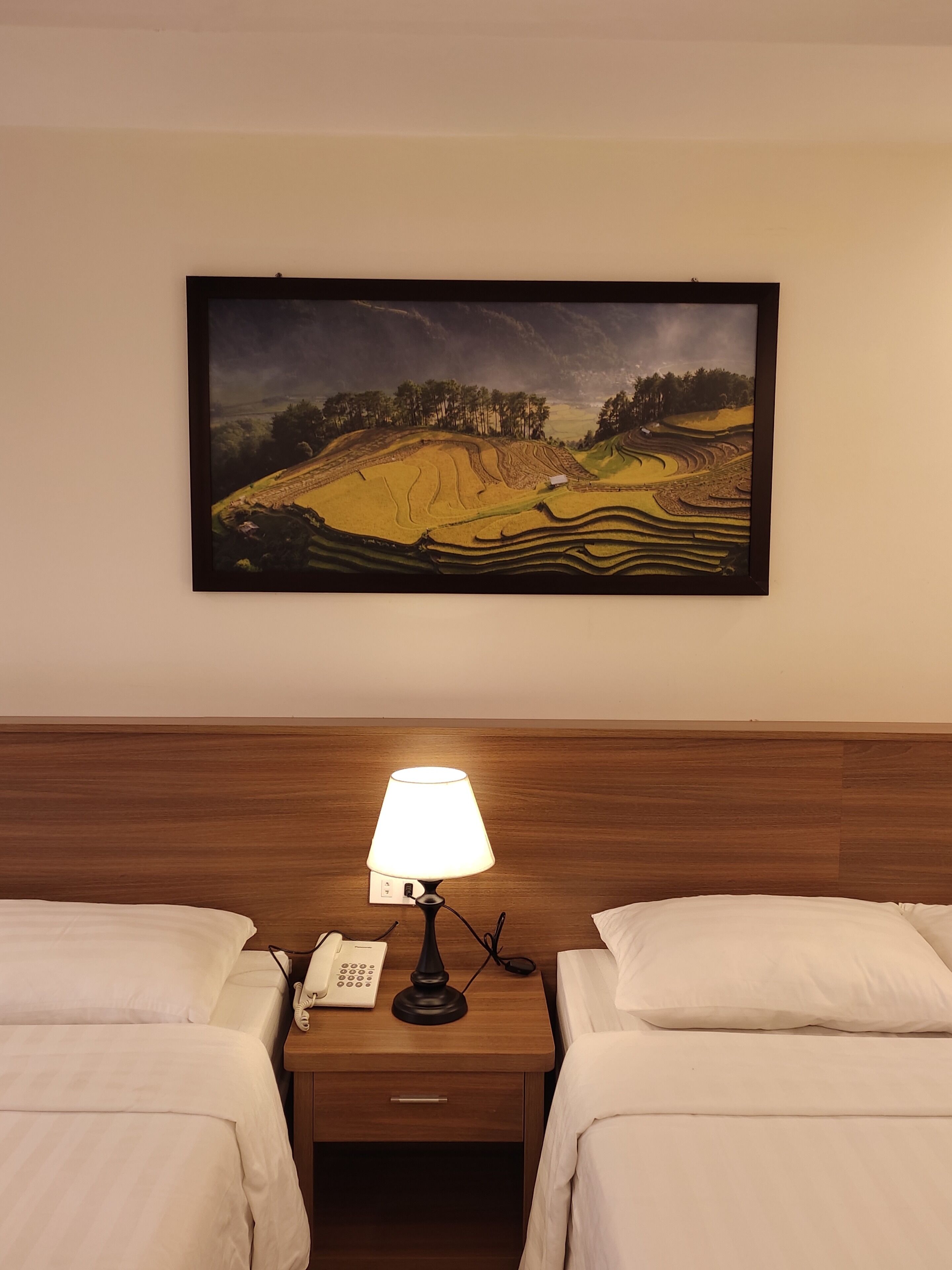 A hotel room with two beds, a nightstand with a lamp, a phone, and a painting of a landscape on the wall.