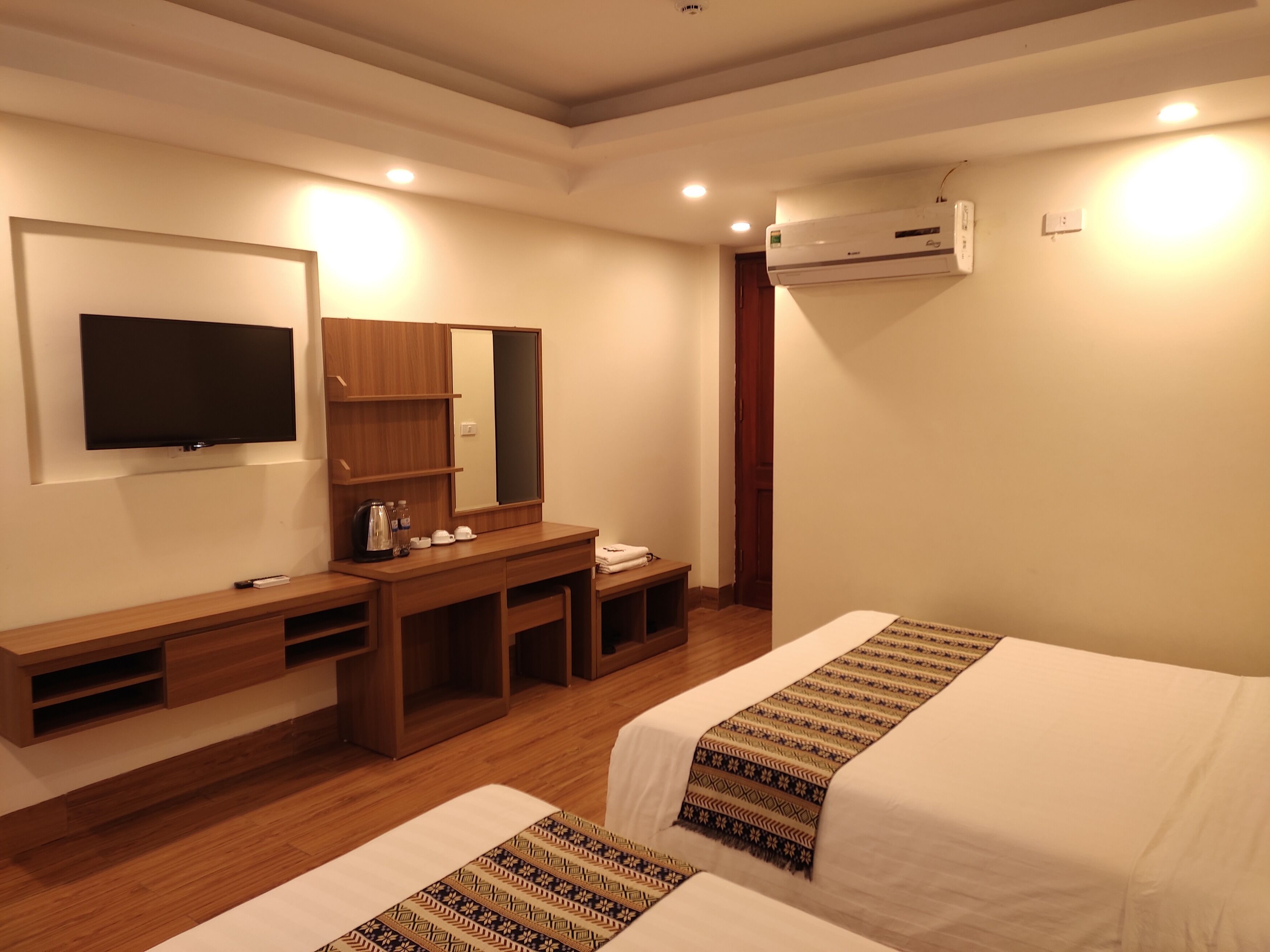 A hotel room with a flat-screen TV, wooden desk, two beds with patterned bedspreads, a mirror, and an air conditioning unit.