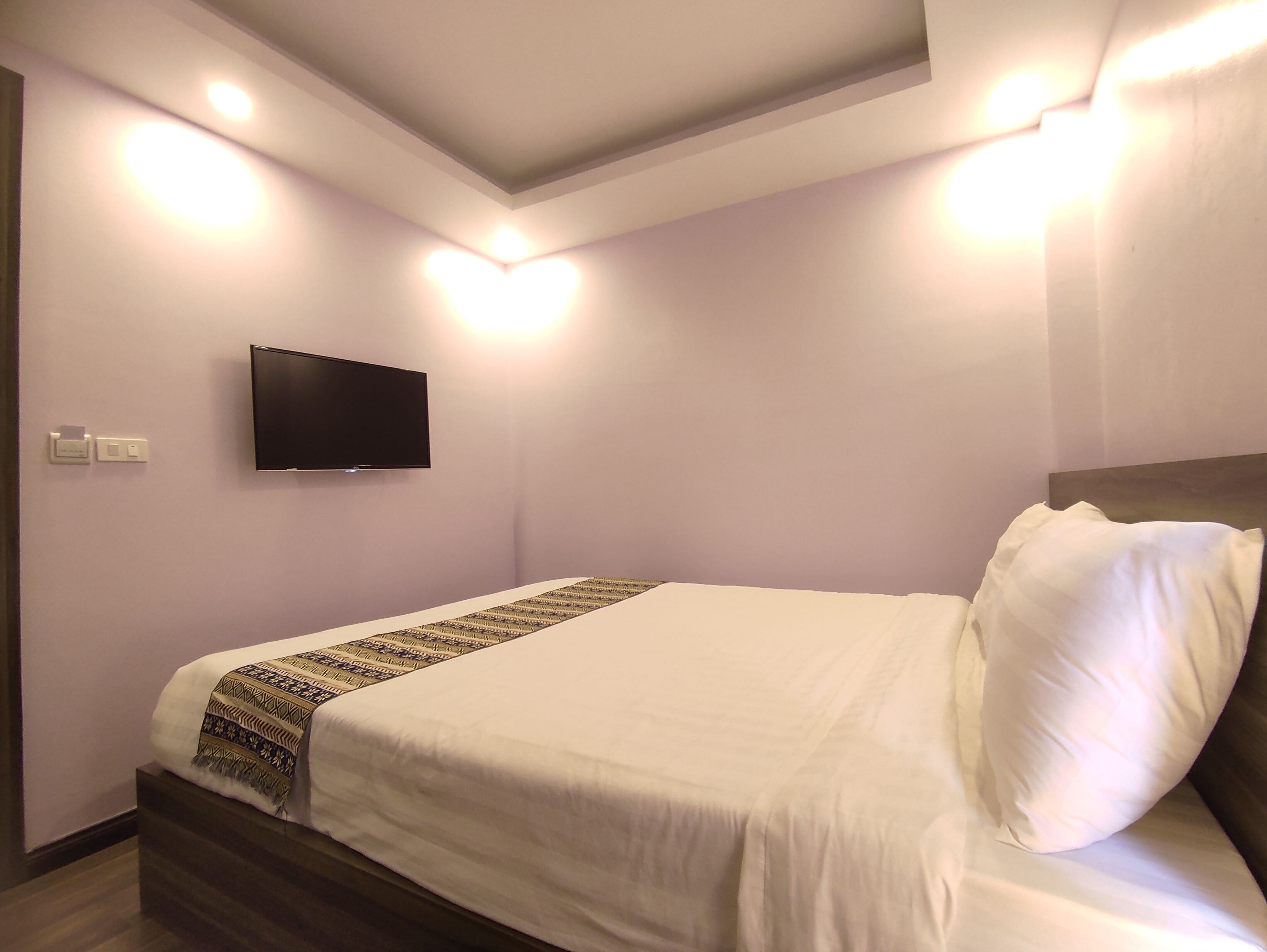 A neatly made bed with a patterned throw, a flat-screen TV mounted on the wall, and recessed ceiling lighting.