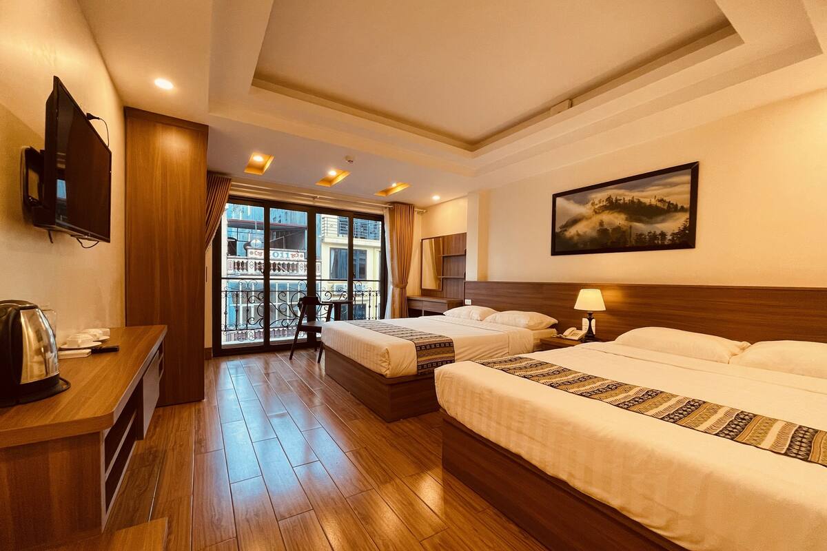 Habitación de hotel con dos camas, un escritorio con hervidor, televisión y balcón con vistas a la ciudad.