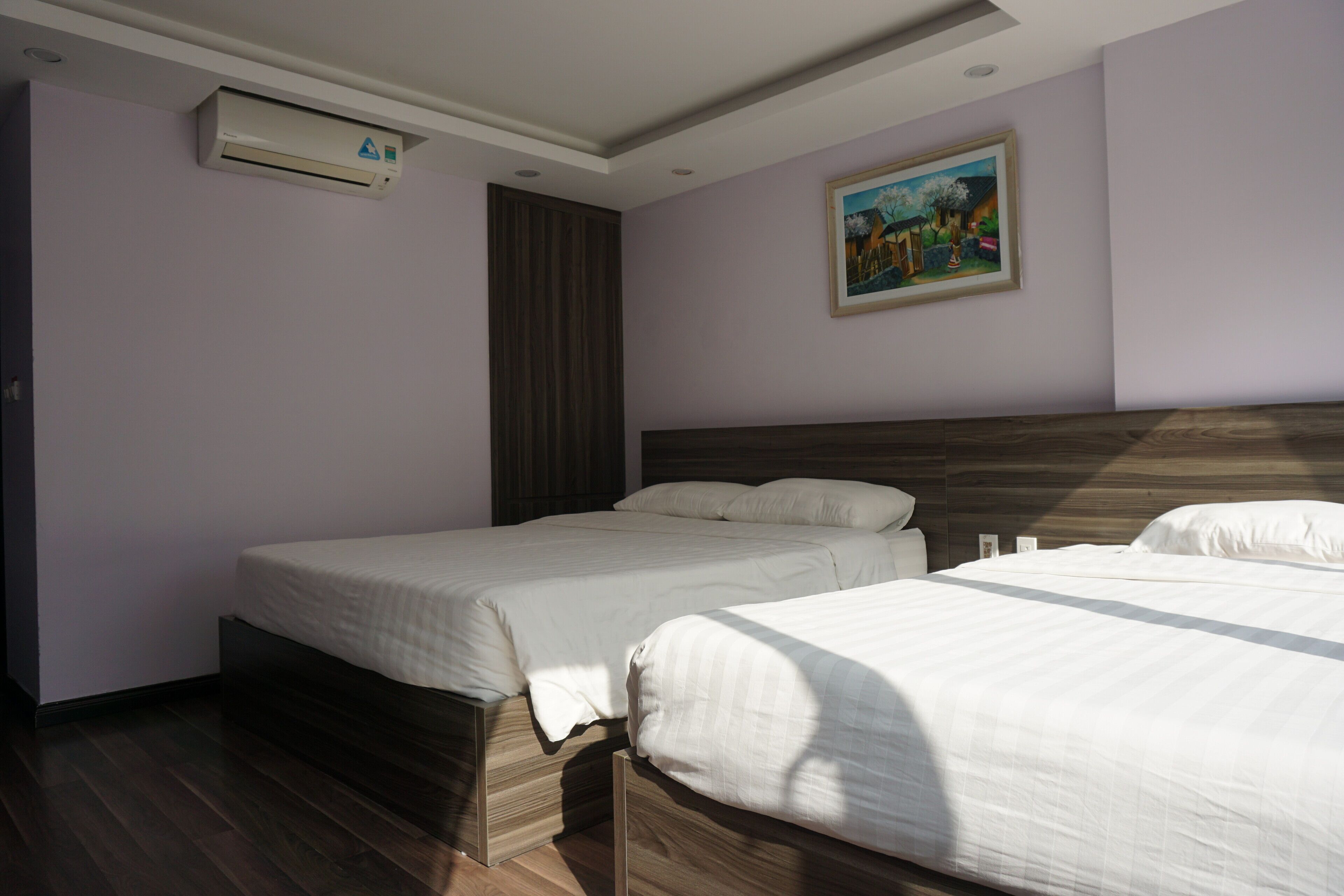 A bedroom with two beds, wooden headboards, a ceiling-mounted air conditioner, and a framed painting on the wall.
