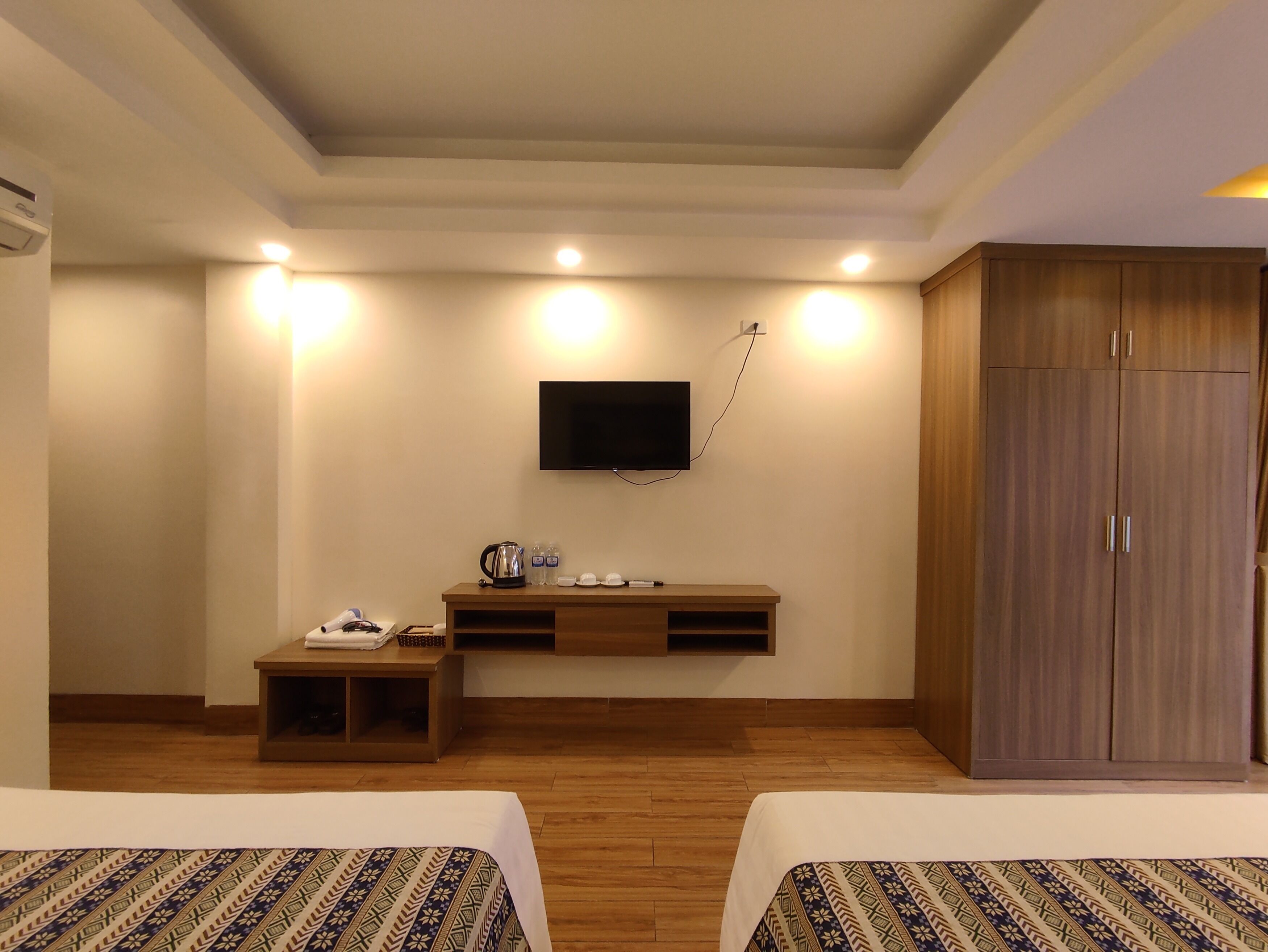 A hotel room with two beds, a wooden cabinet, a TV mounted on the wall, and a small table with a kettle and cups.