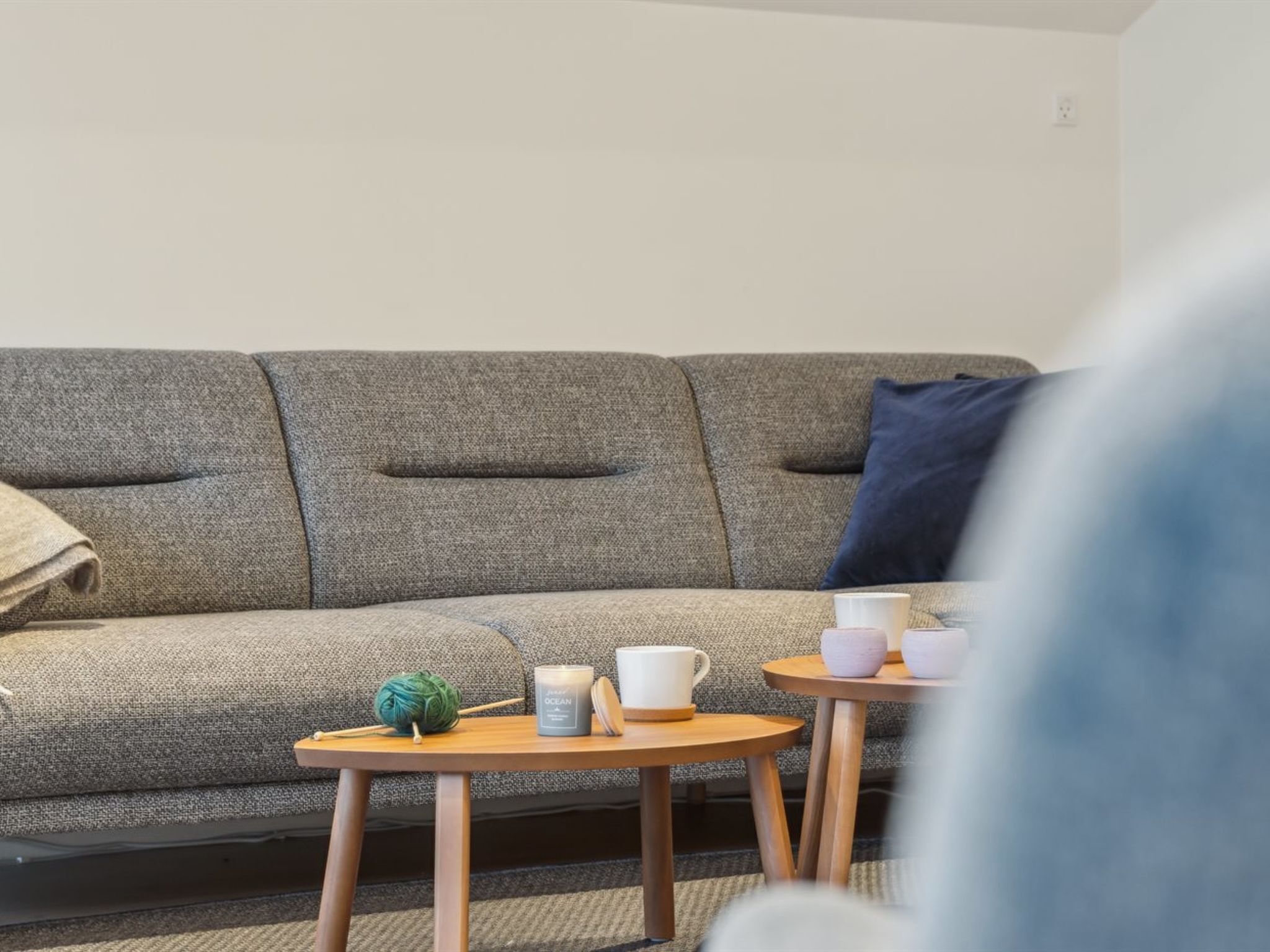 A grey sofa with a blue pillow, a small wooden table with a candle, a ball of yarn, and two cups.