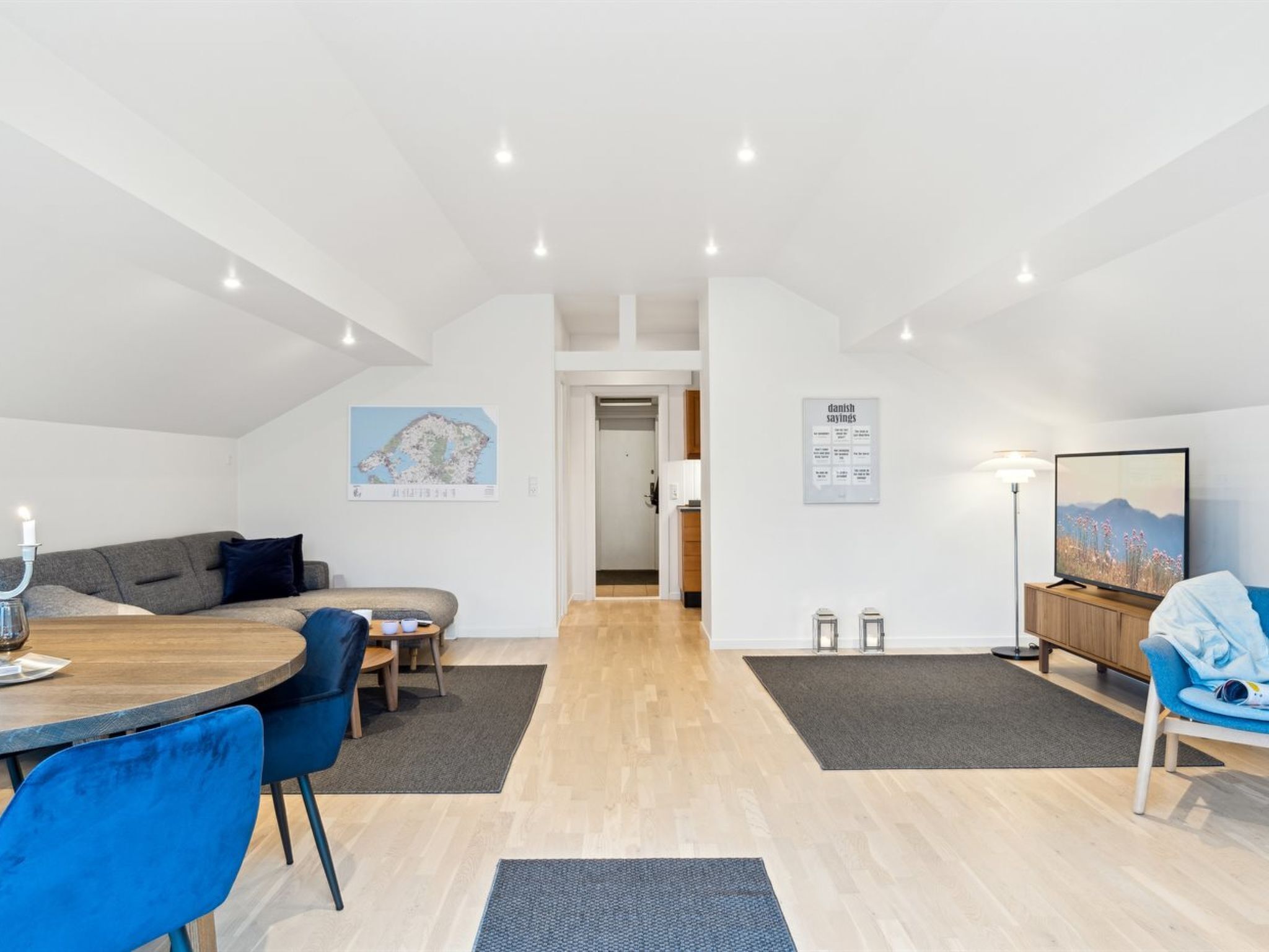 A modern, well-lit room with a wooden dining table, blue chairs, a sofa, and a TV.