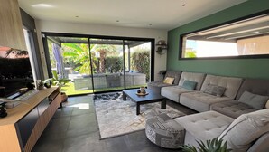 A modern living room with a sofa, coffee table, and TV stand. There is a sliding glass door leading to a balcony with plants. - Villa with swimming pool and Jacuzzi, ideal for families, beach 10 min away, residential (La Crau)