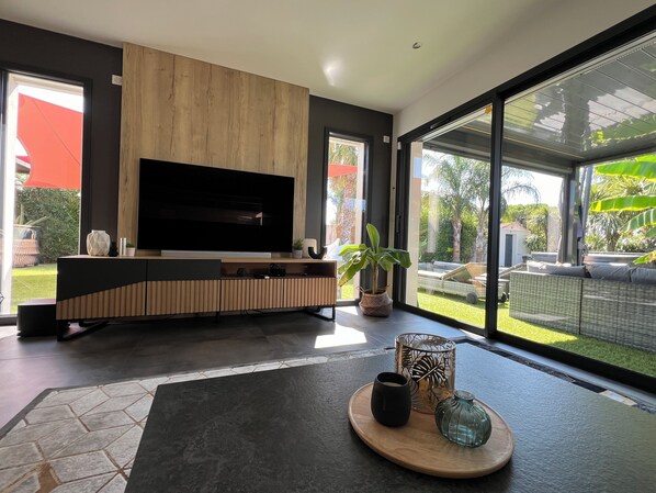 A modern living room with a large flat-screen TV, wooden paneling, a potted plant, and a view of a garden through large glass doors.