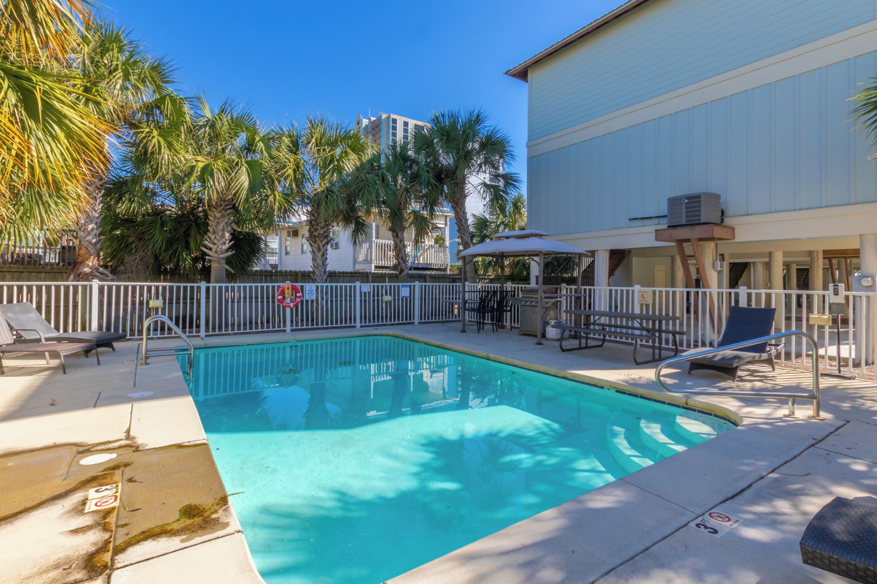 A rectangular swimming pool with a white fence, lounge chairs, and a covered seating area with umbrellas.