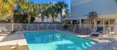 A rectangular swimming pool with a white fence, lounge chairs, and a covered seating area with umbrellas.