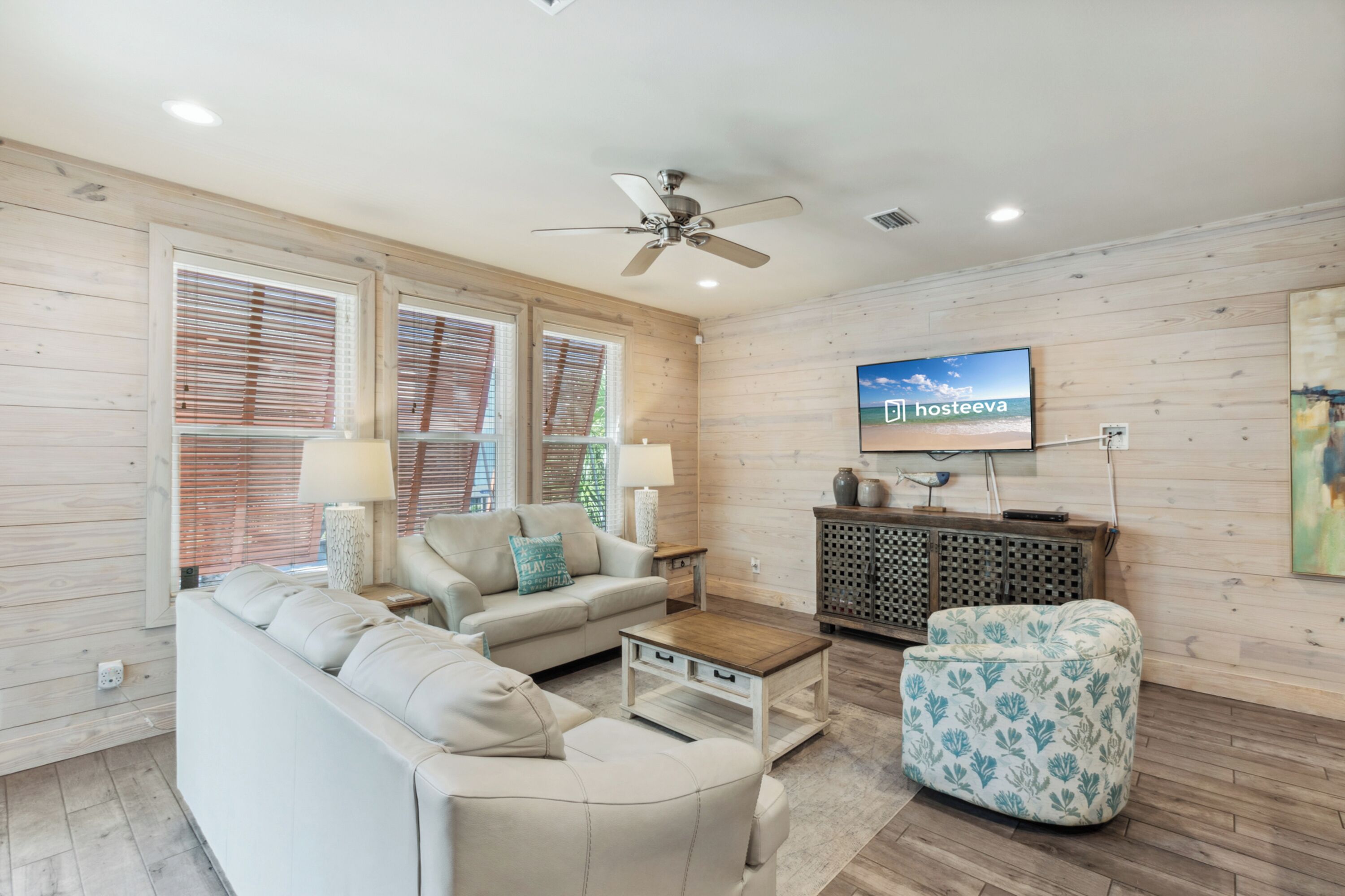 A modern living room with a sofa, coffee table, and ottoman, a ceiling fan, and a television mounted on the wall.