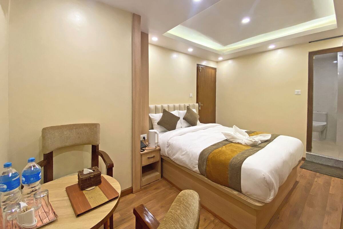 A hotel room with a bed, bedside table, chair, and a small table with water bottles and a tray.