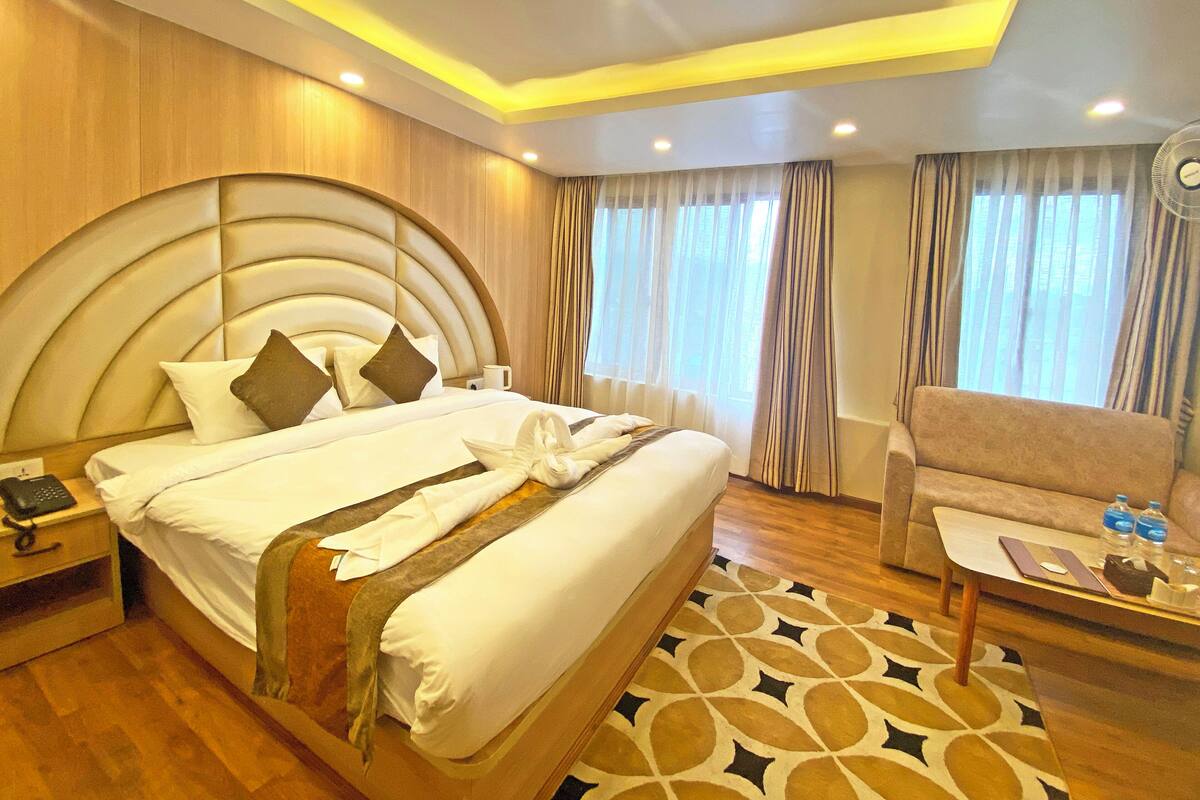 A hotel room with a large bed, a bedside table with a phone, a chair, and a small table with a fan and water bottles.