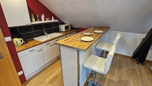 A compact kitchen with a wooden countertop, white cabinets, a microwave, and a sink. There are two white bar stools and a small shelf with decorative items.