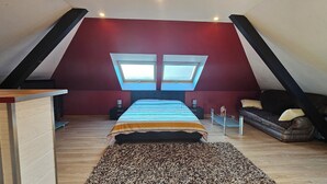 A bedroom with a sloped ceiling, two skylights, a bed, a nightstand, a small table, a sofa, and a patterned rug.