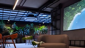 A modern interior with a glass vase on a wooden table, a brown sofa, and a large screen displaying a nature scene. - Way Tambaú  (João Pessoa)