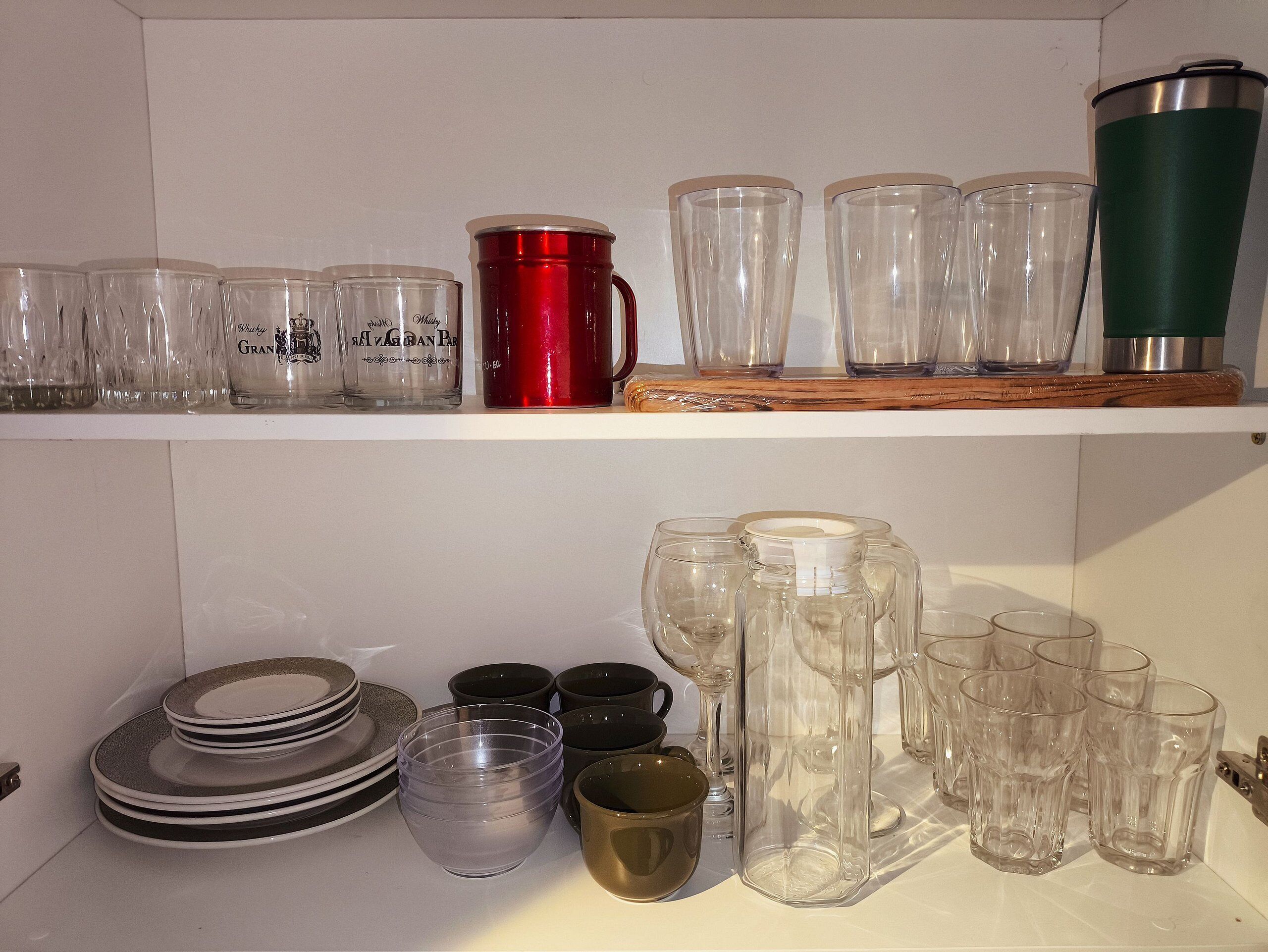 A shelf with various glassware including tumblers, a red mug, and a green tumbler. Below, there are stacked plates and bowls.