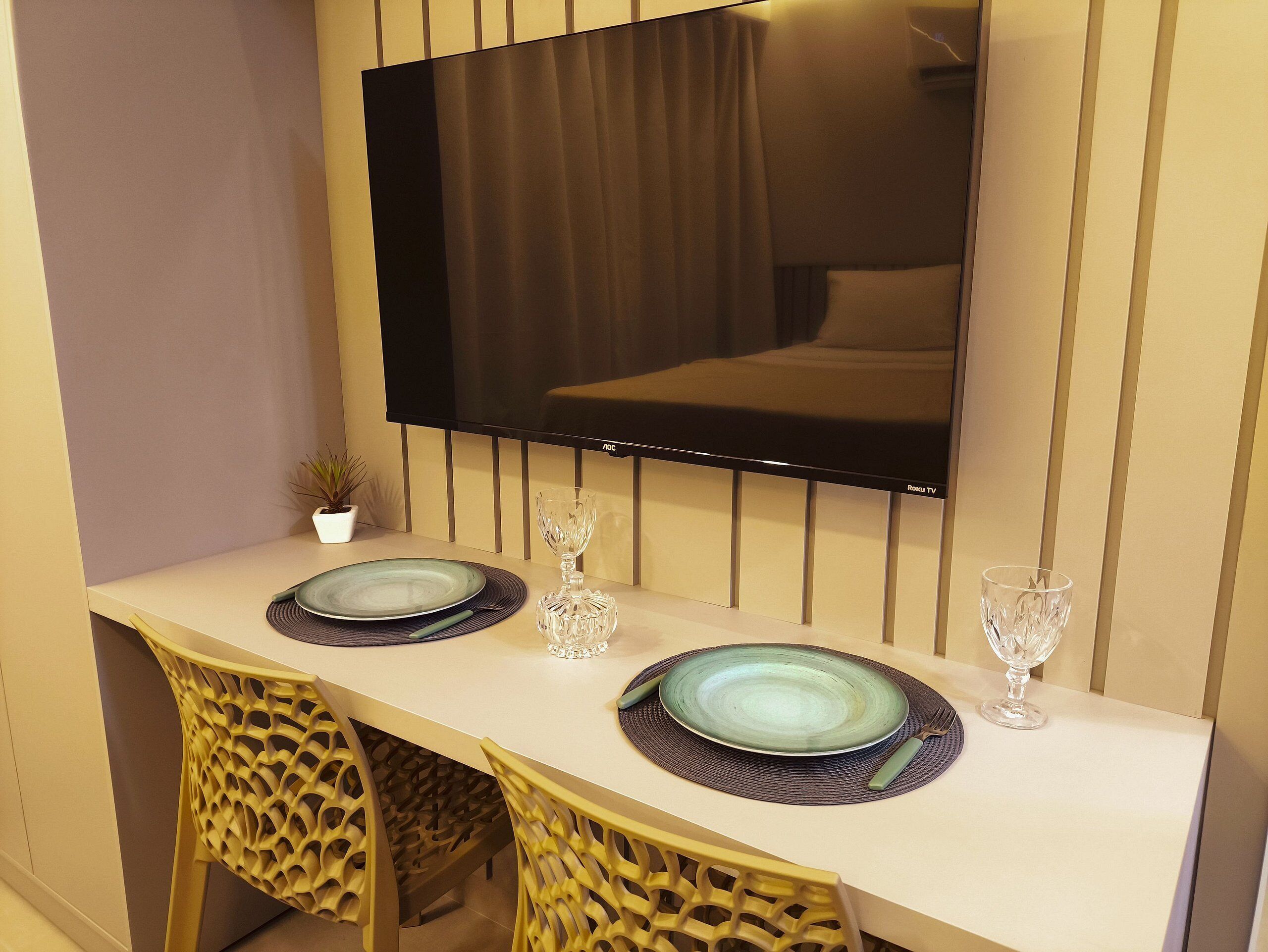 A dining area with a table set for two, a television mounted on the wall, and a small potted plant.