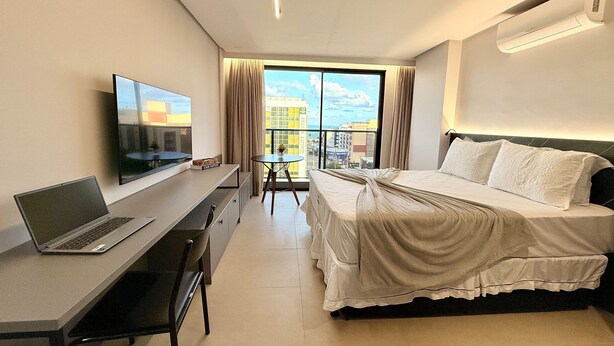 A hotel room with a bed, a desk with a laptop, a TV, and a view of the city through large windows. - Way Tambaú  (João Pessoa)